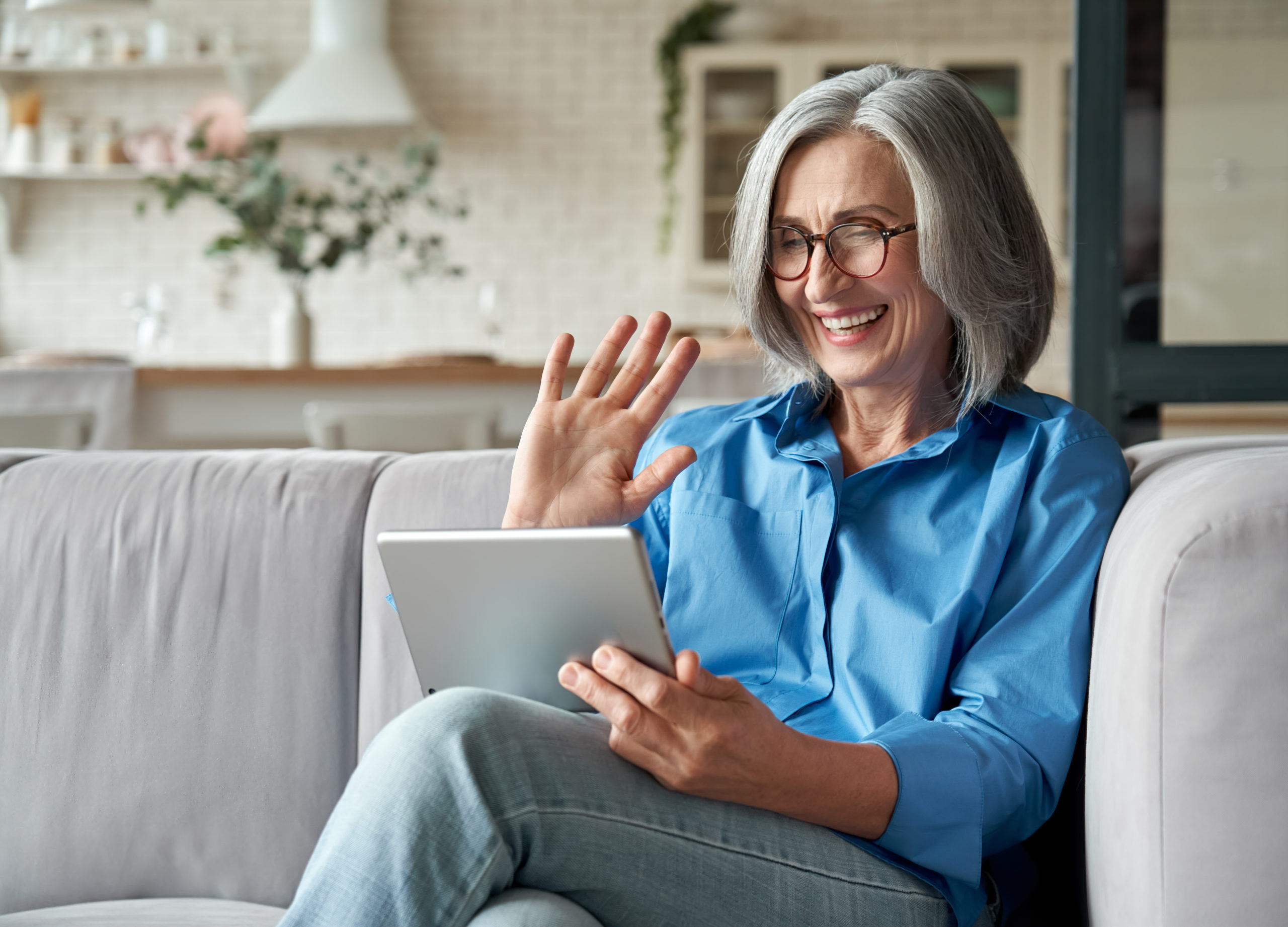 Happy 60s older mature middle aged adult woman waving hand holding digital tablet computer video conference calling by social distance virtual family online chat meeting sitting on couch at home.
