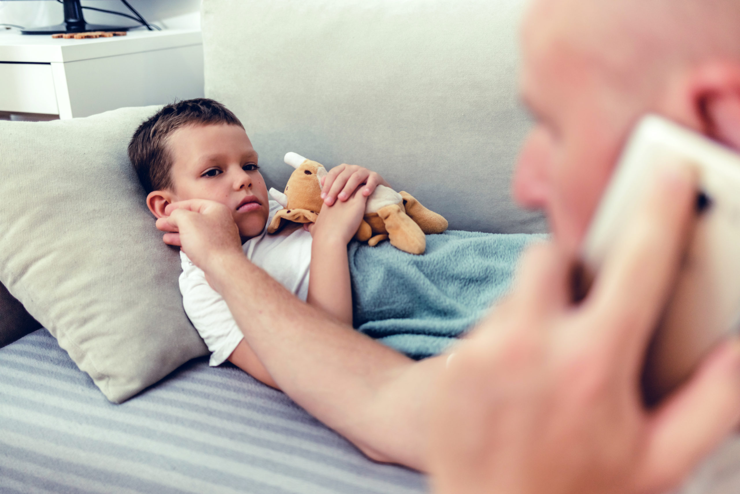 Worried father consulting doctor on cellphone while sitting next to his little son in the living room.